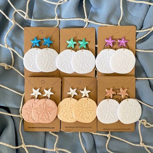 Pode incluir: Seis pares de brincos de d&oacute;lar de areia em papel&atilde;o marrom. Cada par apresenta um d&oacute;lar de areia branco com um design em relevo e um amuleto de estrela do mar em v&aacute;rias cores, incluindo rosa, azul, turquesa e branco.