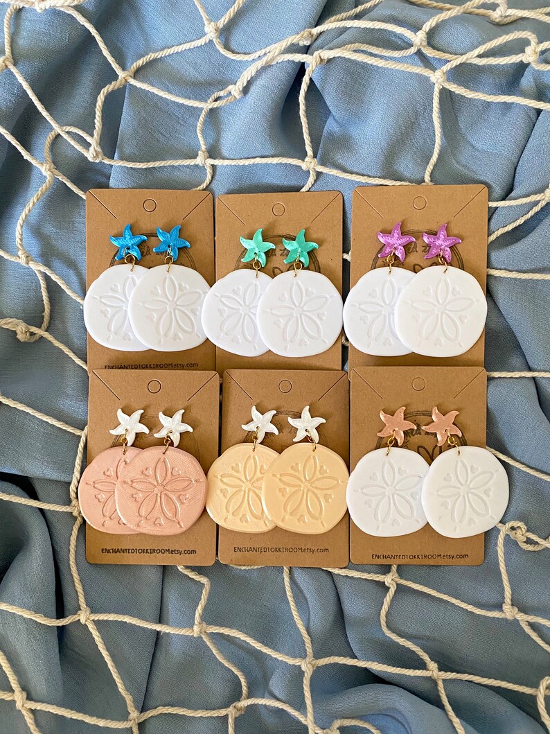 Pode incluir: Seis pares de brincos de d&oacute;lar de areia em papel&atilde;o marrom. Cada par apresenta um d&oacute;lar de areia branco com um design em relevo e um amuleto de estrela do mar em v&aacute;rias cores, incluindo rosa, azul, turquesa e branco.