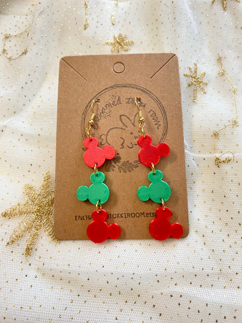 May include: Red and green Mickey Mouse-shaped dangle earrings with gold hooks. The earrings are on a brown card with the text "Enchanted Room Etsy".