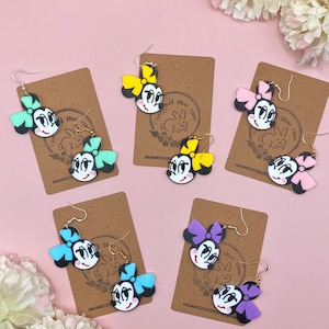 May include: Four pairs of dangle earrings featuring a cartoon character with a bow. The earrings are on brown cards with the text "Enchanted Dixie Designs". The bows are in different colours: yellow, pink, blue, and purple.