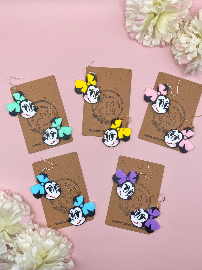 May include: Four pairs of dangle earrings featuring a cartoon character with a bow. The earrings are on brown cards with the text "Enchanted Dixie Designs". The bows are in different colours: yellow, pink, blue, and purple.