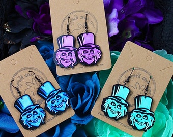 May include: Three pairs of dangle earrings featuring a ghost character with a top hat. The earrings are made of wood and have a black outline. The ghosts are painted in purple, blue, and green.