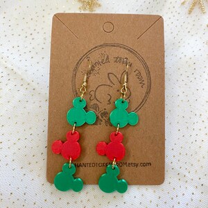 May include: A pair of dangle earrings featuring three green and two red Mickey Mouse head charms. The earrings are on a brown card with the text "Haunted10kroom.etsy.com".