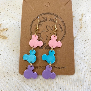 May include: A pair of dangle earrings with three layered charms shaped like Mickey Mouse heads. The charms are pink, blue, and purple.