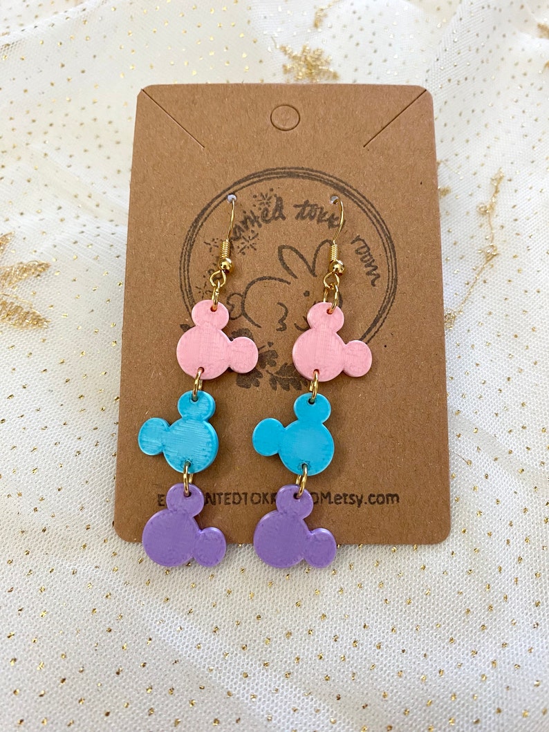 May include: A pair of dangle earrings with three layered charms shaped like Mickey Mouse heads. The charms are pink, blue, and purple.