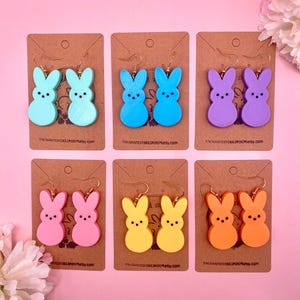 May include: Six pairs of pastel-colored bunny-shaped earrings. The earrings are made of wood and have gold hooks. The colors are light blue, blue, purple, pink, yellow, and orange.