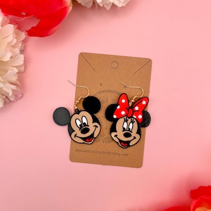May include: A pair of dangle earrings featuring Mickey Mouse and Minnie Mouse. The earrings are made of black, red, and white plastic and have gold hooks. The earrings are on a brown card with the text "ENCHANTEDICKERBROOM.etsy.com".