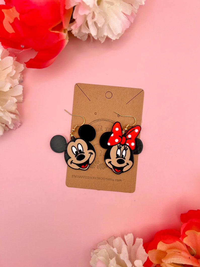 May include: A pair of dangle earrings featuring Mickey Mouse and Minnie Mouse. The earrings are made of black, red, and white plastic and have gold hooks. The earrings are on a brown card with the text "ENCHANTEDICKERBROOM.etsy.com".