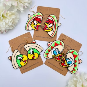 Könnte beinhalten: Ein Paar bunte Comic-Figuren-Ohrringe mit Sombrero-Design. Die Ohrringe sind aus Acryl und haben ein grün-rot-gelbes Farbschema. Die Ohrringe hängen an goldenen Haken.