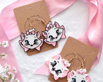May include: A pair of white and pink cat-shaped earrings with gold hoops. The earrings feature a cartoon-style cat face with pink bows and black eyes. The earrings are displayed on a brown card with the text "EnchantedTokiRoom" and a website address.