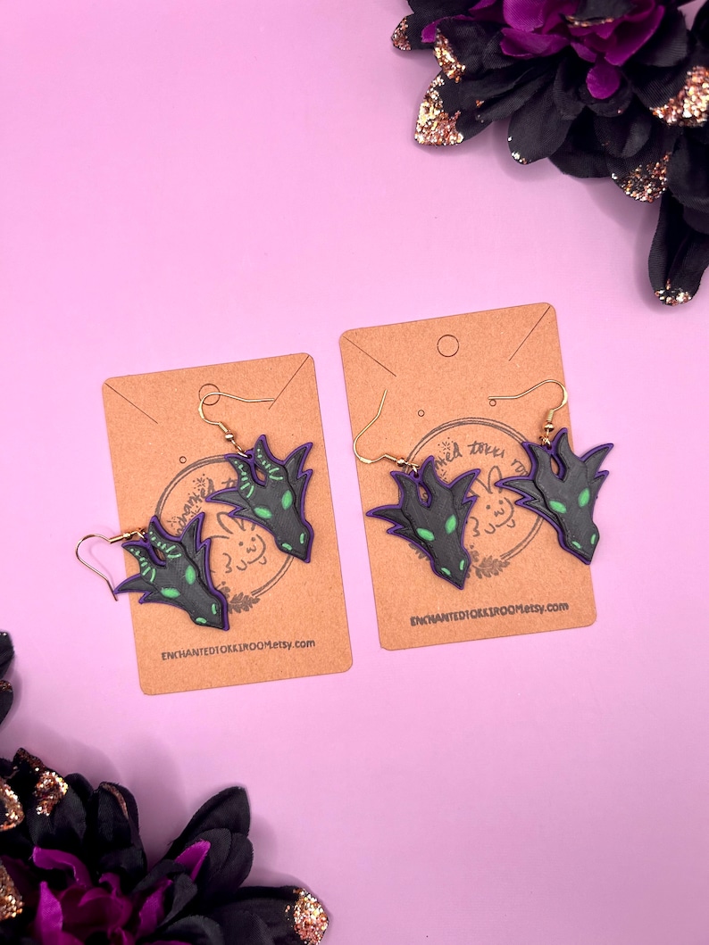May include: A pair of dangle earrings featuring a black and green dragon head design. The earrings are on a brown card with the text "ENCHANTEDICKOOKROOM.etsy.com".