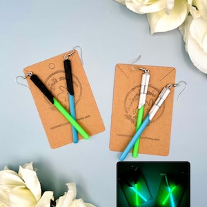 May include: Two pairs of glow-in-the-dark lightsaber earrings. The earrings are made of black, green, and blue polymer clay. The earrings are shown on a brown card with the text "ENCHANTED DREAMS" and "ROOM.etsy.com". The earrings are also shown glowing in the dark.