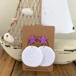 Pode incluir: Um par de brincos de d&oacute;lar de areia brancos com pingentes de estrela do mar roxos. Os brincos est&atilde;o em um cart&atilde;o marrom com o texto "enchantedokkiroom.etsy.com".