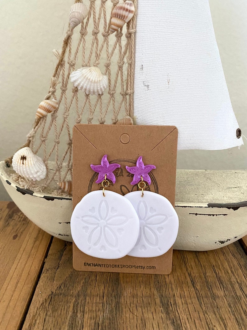 Pode incluir: Um par de brincos de d&oacute;lar de areia brancos com pingentes de estrela do mar roxos. Os brincos est&atilde;o em um cart&atilde;o marrom com o texto "enchantedokkiroom.etsy.com".