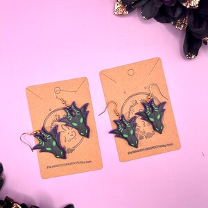 May include: A pair of dangle earrings featuring a black and green dragon claw design with purple accents. The earrings are displayed on brown card stock with the Etsy shop name "enchantedoxxbroom.etsy.com" printed on each card.