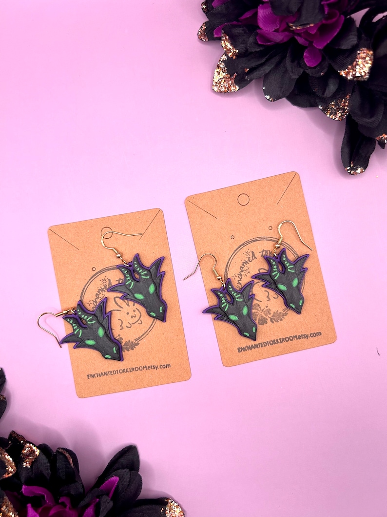 May include: A pair of dangle earrings featuring a black and green dragon claw design with purple accents. The earrings are displayed on brown card stock with the Etsy shop name "enchantedoxxbroom.etsy.com" printed on each card.