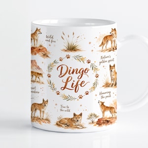 Può includere: Una tazza in ceramica bianca con illustrazioni ad acquerello di dingo in varie pose, piante del deserto e impronte di zampe. Il testo "Dingo Life" è centrato all'interno di una ghirlanda botanica, accompagnato da frasi come "Wild and free" e "Nature's golden spirit."