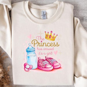 Puede incluir: Una sudadera de color crema con el texto "The Princess has arrived it's a girl!" en letras rosas y doradas. Una corona, un biberón y zapatos de bebé están ilustrados en la sudadera.