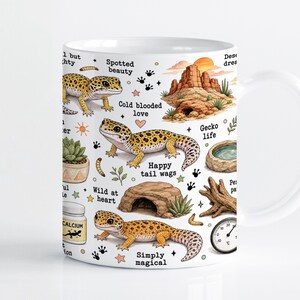 Könnte beinhalten: Ein weißer Keramikbecher mit einem wiederkehrenden Muster aus Leopardgeckos, Wüstenfelsformationen, einer Sukkulente, einer Wasserschale, Treibholz, einem Kalziumbehälter und einem Thermometer. Der Text enthält "Spotted beauty", "Cold blooded love" und "Gecko life".