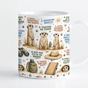Puede incluir: Una taza de cerámica blanca con una ilustración colorida de suricatas, binoculares, un sombrero de sol, una cantimplora y un cuaderno. El diseño incluye frases numeradas como "Small creatures big hearts", "Adventure awaits" y "Keep your eyes on the wonders".