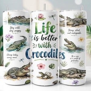 May include: Three white cylindrical tumblers with straws featuring watercolour crocodile illustrations, tropical flowers, and dragonfly motifs. Text includes "Life is better with Crocodiles" and phrases like "Stay wild, stay snappy" and "Chomp today, worry never."
