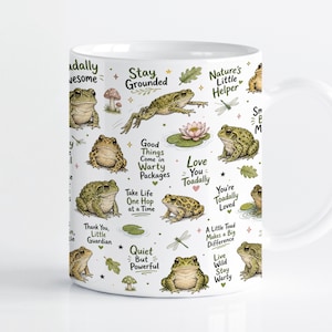 Pode incluir: Uma caneca de cerâmica branca com um padrão repetitivo de sapos verdes, vitórias-régias, libélulas e cogumelos. O design inclui frases como Stay Grounded, Toadally Awesome, Love You Toadally e A Little Toad Makes a Big Difference.