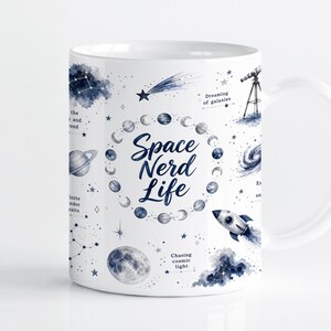 Pode incluir: Uma caneca de cerâmica branca com um padrão de tema espacial em azul escuro com planetas, estrelas, um foguete, um telescópio e uma lua. O texto "Space Nerd Life" está impresso no centro, rodeado por uma disposição circular de fases lunares.