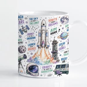 Może przedstawiać: Biały ceramiczny kubek z kolorową ilustracją o tematyce kosmicznej, przedstawiającą startującą rakietę, hełm astronauty, teleskop, planety i gwiazdy. Projekt zawiera motywacyjne napisy, takie jak "Reach for the stars", "Ignite your dreams" oraz "Gravity is just a suggestion" w różnych czcionkach i kolorach.