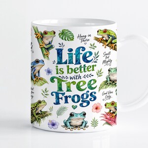 Pode incluir: Uma caneca de cerâmica branca com uma ilustração colorida de várias rãs, folhas verdes e flores tropicais. O texto diz: Life is better with Tree Frogs, Hang in There, Small But Mighty e Good Vibes Only em letras azuis e verdes.