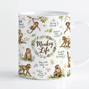 Puede incluir: Una taza de cerámica blanca con un patrón repetitivo de monos marrones en varias posturas, follaje verde y el texto "Monkey Life" en el centro. Las frases adicionales incluyen "Wild heart", "Swing into joy", "Nature's little acrobat" y "Stay wild, little one".