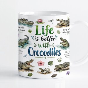 Puede incluir: Una taza de cerámica blanca con una colorida ilustración en acuarela de varios cocodrilos, flores de loto y libélulas. El texto "Life is better with Crocodiles" está impreso en el centro en letras verdes y azules de estilo manuscrito.