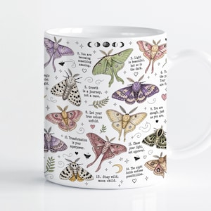 May include: A white ceramic mug featuring a colorful pattern of various moth species in shades of green, purple, pink, and brown. The design includes small stars, moon phases, and inspirational text phrases such as 'Transformation is your superpower' and 'Stay wild, moon child' scattered among the insects.