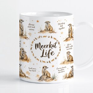 Pode incluir: Uma caneca de cerâmica branca com um padrão de ilustrações de suricatas castanhas, pequenas estrelas douradas e pegadas. O texto central diz Meerkat Life, rodeado por frases como Always alert and curious, Standing tall together e Life is better with meerkats.
