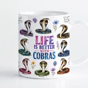 Puede incluir: Una taza de cerámica blanca con un estampado estilo acuarela de varias cobras en diferentes poses y colores. El texto central dice "LIFE IS BETTER WITH COBRAS" en letras negritas multicolores, rodeado de pequeñas ilustraciones de flores.