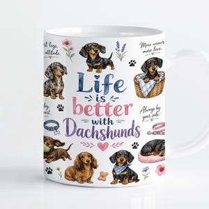Puede incluir: Una taza de cerámica blanca con ilustraciones en acuarela de varios perros salchicha en diferentes posturas, incluyendo sentados, tumbados y dentro de una cesta. El texto dice Life is better with Dachshunds, rodeado de flores y huellas.