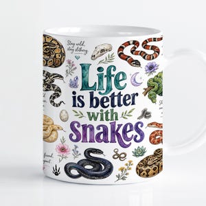 Puede incluir: Una taza de cerámica blanca con una colorida ilustración en acuarela de varias especies de serpientes, un cráneo, flores y cristales. El texto "Life is better with Snakes" está impreso en el centro en azul y morado, con "Stay wild, stay slithery" arriba.
