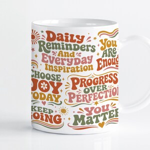 May include: A white ceramic mug featuring a multi-coloured graphic print with the text Daily Reminders And Everyday Inspiration, You Are Enough, Choose Joy Today, Progress Over Perfection, Keep Going, and You Matter, accented by small suns and hearts.
