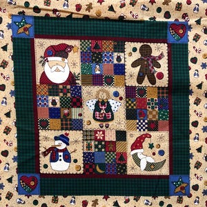 May include: Festive Christmas fabric with a patchwork design. The central panel features Santa Claus, a gingerbread man, an angel, a snowman, and a crescent moon with a star. The fabric has a beige background with festive icons.
