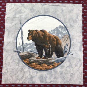 May include: A square fabric panel featuring a central circular illustration of a brown bear standing on a rock in a mountainous landscape. The border is a deep maroon color with a repeating pattern of small light blue wolf silhouettes.