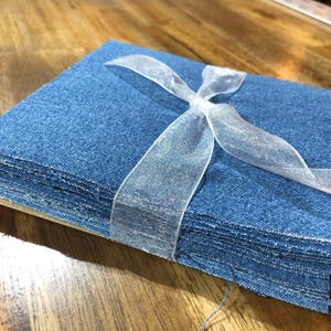 May include: A stack of rectangular blue denim fabric swatches tied together with a translucent white organza ribbon. The fabric has a visible woven texture and frayed edges, secured in a cross pattern by the sheer ribbon tied in a knot.