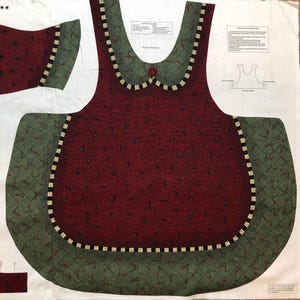 May include: A printed fabric panel for a sewing project featuring a dark red apron body with a black crackle pattern and small black teardrop shapes. The apron has a green collar and hem with a holly leaf pattern and a black and white checkered trim.