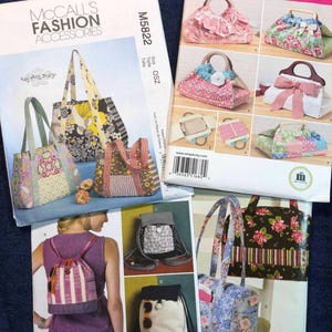 May include: Several sewing pattern packets for bags and backpacks are displayed on a blue surface. The packets feature illustrations of various bag designs, including tote bags and backpacks, with floral and patterned fabrics. The McCall's and Simplicity brands are visible.