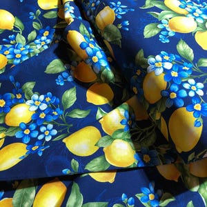 May include: A close-up of a fabric with a repeating pattern of yellow lemons, blue flowers, and green leaves on a dark blue background. The fabric is folded, showing the pattern in multiple layers. The design is bright and cheerful.