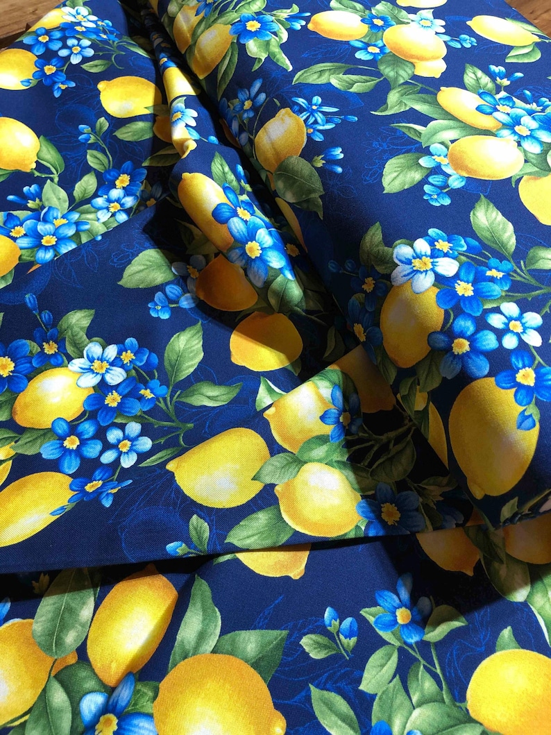 May include: A close-up of a fabric with a repeating pattern of yellow lemons, blue flowers, and green leaves on a dark blue background. The fabric is folded, showing the pattern in multiple layers. The design is bright and cheerful.