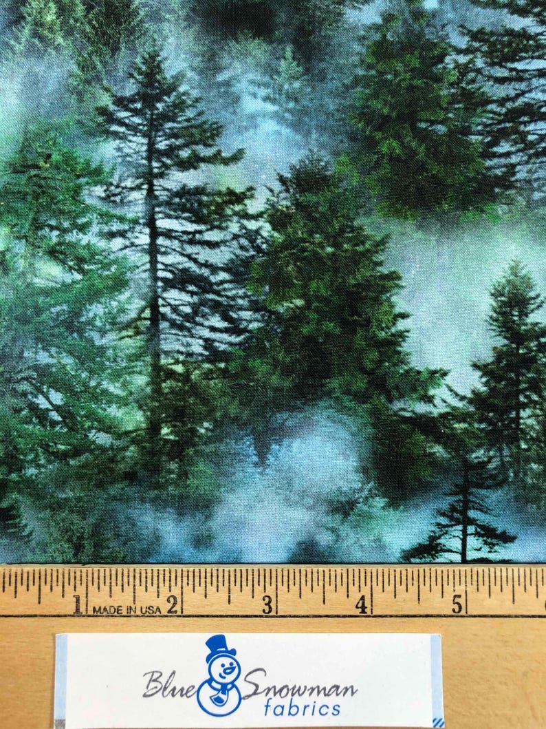 Op de afbeelding: Stof met een boslandschap met hoge groene dennenbomen en een mistige blauwe achtergrond. Langs de onderkant is een liniaal te zien en een label met "Blue Snowman fabrics".