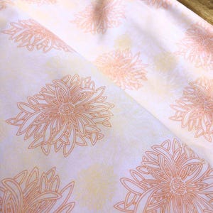 Può includere: Primo piano di tessuto bianco con un motivo ripetuto di grandi disegni floreali arancione-marrone. I fiori hanno contorni dettagliati e sono posti su uno sfondo giallo chiaro sottile. Il tessuto è piegato, mostrando la sua consistenza.