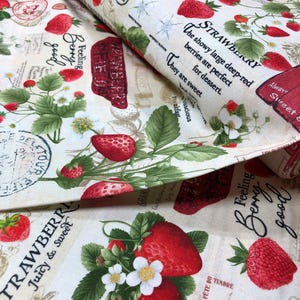 May include: A white fabric with a repeating pattern of red strawberries, green leaves, and white flowers. The fabric has text that says "Feeling Berry good" and "Strawberry Juicy & Sweet".
