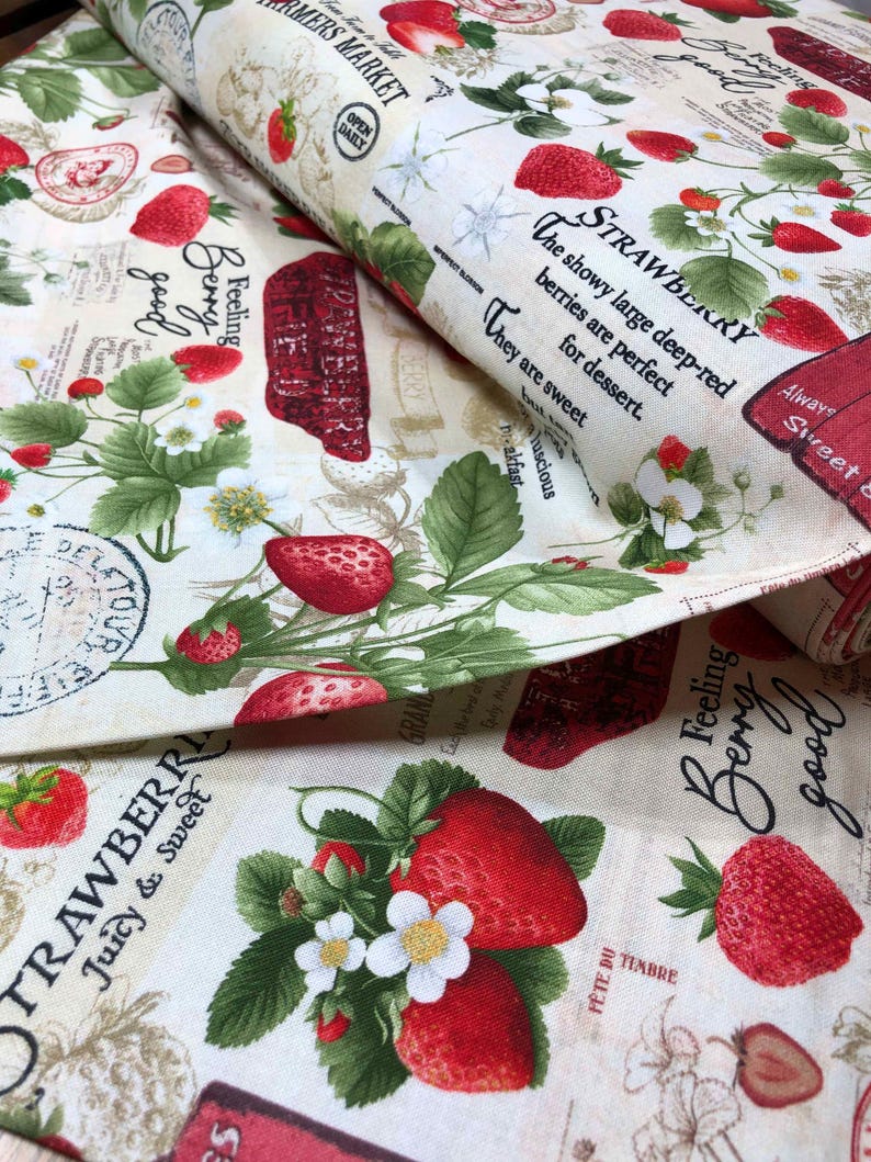 May include: A white fabric with a repeating pattern of red strawberries, green leaves, and white flowers. The fabric has text that says "Feeling Berry good" and "Strawberry Juicy & Sweet".