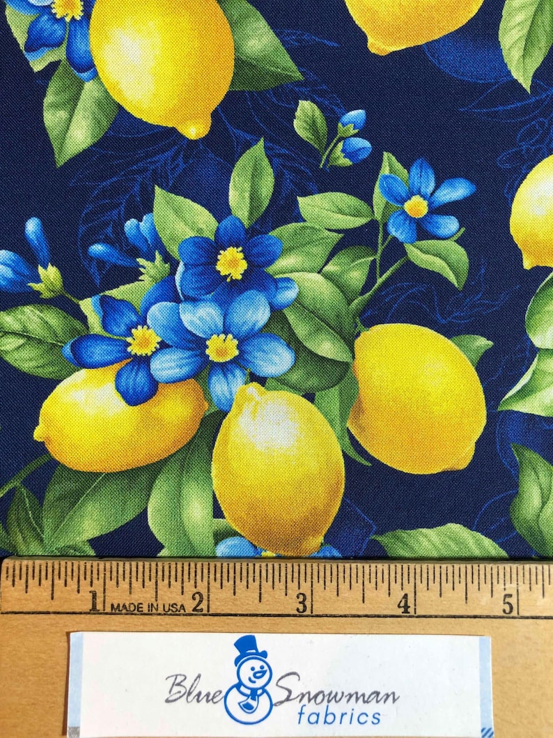 May include: Fabric with a vibrant pattern of yellow lemons, blue flowers, and green leaves on a dark blue background. A wooden ruler with inch measurements is at the bottom. The text "Blue Snowman fabrics" is visible.