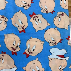 May include: A light blue fabric featuring a repeating pattern of cartoon pig heads. Each pig has pink skin, large eyes, and a small red bow tie. Some pigs are wearing white hats, while others are shown without headwear, all arranged in a scattered layout.